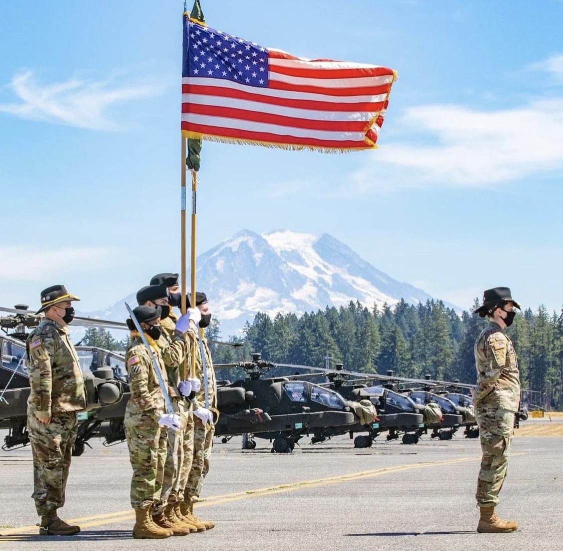 U.S. Army color guard carrying the American flag on a flight line with Apache helicopters and a mountain in the background